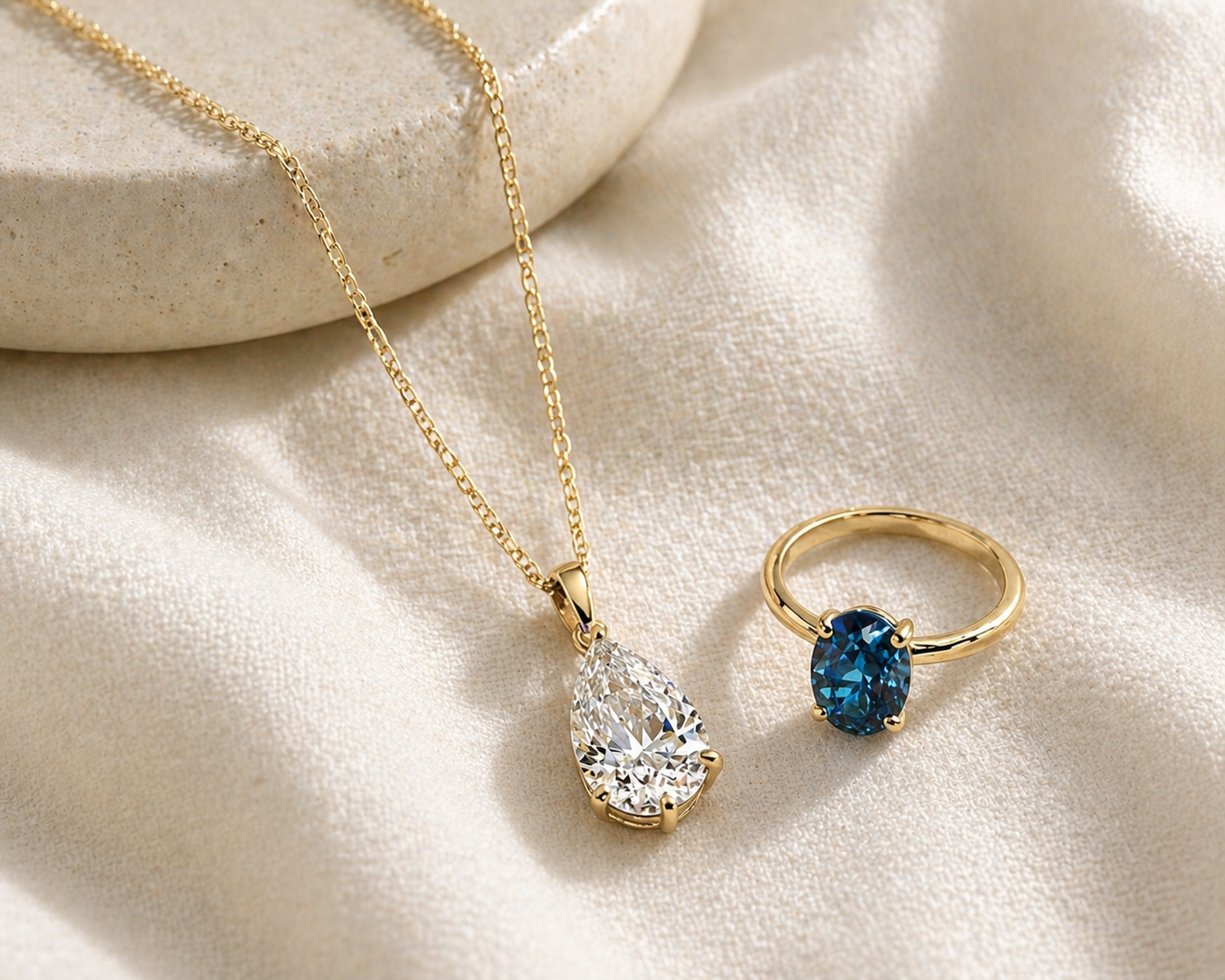 Gold necklace with teardrop-shaped pendant and gold ring with blue lab-grown diamond on a beige fabric background