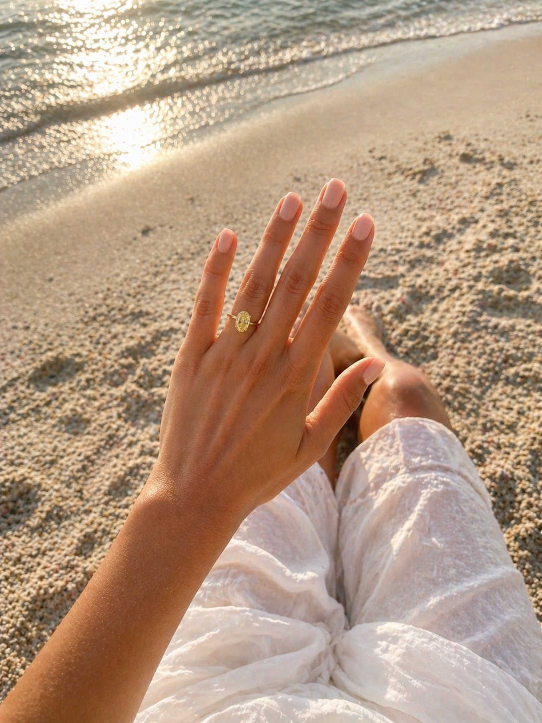 Hand wearing a gold ring on a sandy beach with sunlight reflecting off the water.